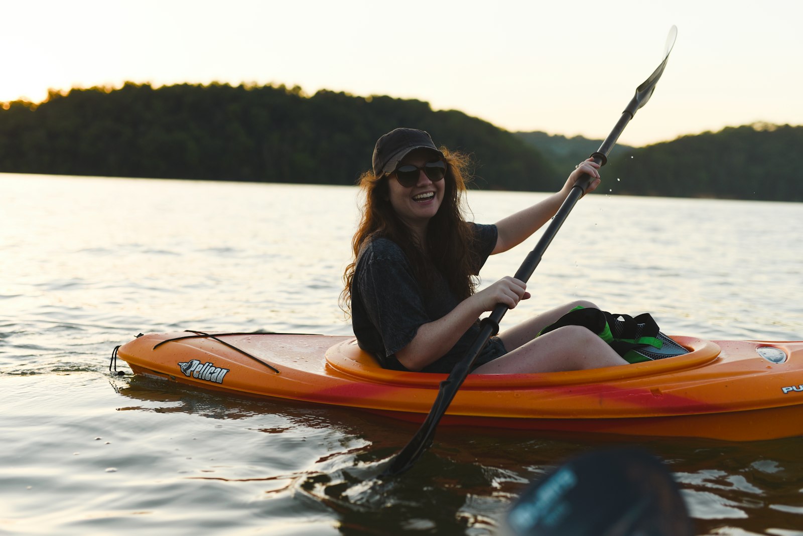 Smiling woman paddling a kayak on a calm lake surrounded by forested hills