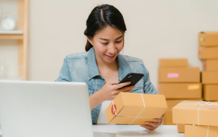 Small business owner checking a holiday order on her phone next to a laptop and stacked shipping boxes