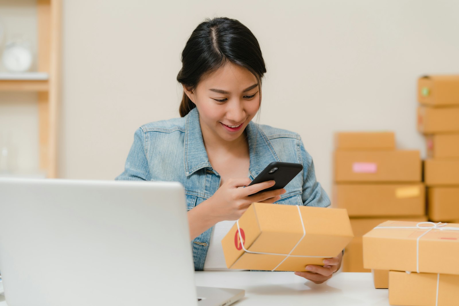 Small business owner checking a holiday order on her phone next to a laptop and stacked shipping boxes
