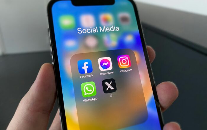 Hand holding a smartphone showing a Social Media folder with Facebook, Messenger, Instagram, WhatsApp and X apps