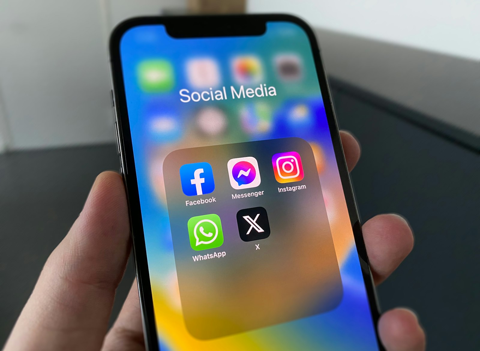Hand holding a smartphone showing a Social Media folder with Facebook, Messenger, Instagram, WhatsApp and X apps